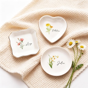 Peut inclure: Trois petits plats à bijoux en céramique blanche avec des bords dorés, chacun avec un motif floral et un nom. Un est carré, un en forme de cœur et un rond. Les noms "Lucy", "Natalia" et "Julie" sont écrits en script. Des marguerites sont placées près des plats.