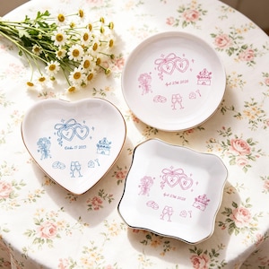 Puede incluir: Tres platos de cerámica blanca con bordes dorados, cada uno con una forma diferente: redondo, corazón y cuadrado. Cada plato presenta un diseño único de boda en rosa o azul, incluyendo corazones, flores y fechas.