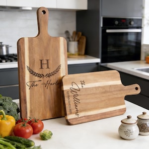 Può includere: Due taglieri in legno con manici. La tavola più grande ha la lettera "H" con una corona d'alloro e il nome "The Harley" inciso. La tavola più piccola ha il nome "The Williams" e "ESTABLISHED 2021" inciso. Verdure e spezie in primo piano.