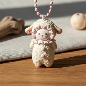 Könnte beinhalten: Eine cremefarbene Schaf-Figur mit rosa Kragen und Ohren, die an einer rosa Perlenkette hängt. Das Schaf hat ein lächelndes Gesicht und steht auf einer Holzoberfläche. Zwei Holzkugeln und ein Stück Treibholz befinden sich im Hintergrund.