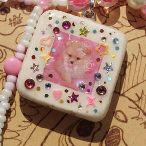 May include: A handcrafted necklace with a square pendant displaying a dog image, embellished with colourful gems and star-shaped details. The necklace features white and pink beaded strands with clear and pink beads.