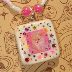 May include: A handmade necklace with a square pendant displaying a dog image, embellished with colourful glitter and star-shaped details. The beaded chain features pink, white, and clear beads.
