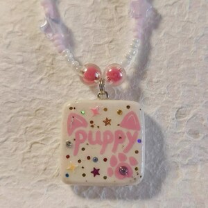 May include: A handmade necklace with a square pendant displaying the word "puppy" in pink, decorated with paw prints, stars, and glitter. The necklace has a beaded chain with pink and white beads.