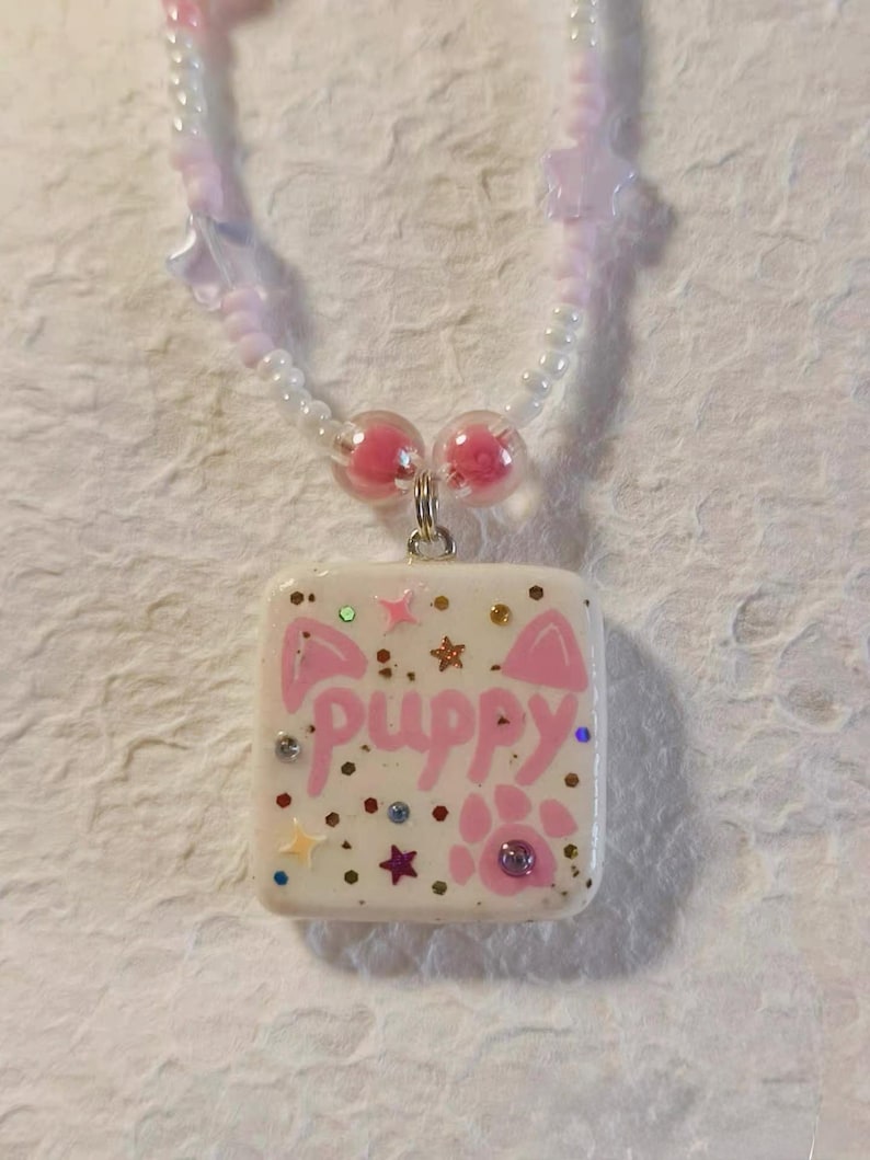 May include: A handmade necklace with a square pendant displaying the word "puppy" in pink, decorated with paw prints, stars, and glitter. The necklace has a beaded chain with pink and white beads.