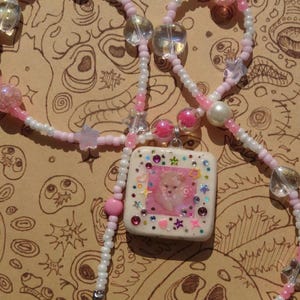 May include: A handcrafted necklace with a square pendant featuring a pink and white design and a small dog image. The necklace has a beaded chain in white and pink, with clear and pink beads. The pendant is decorated with colourful gems and star shapes.