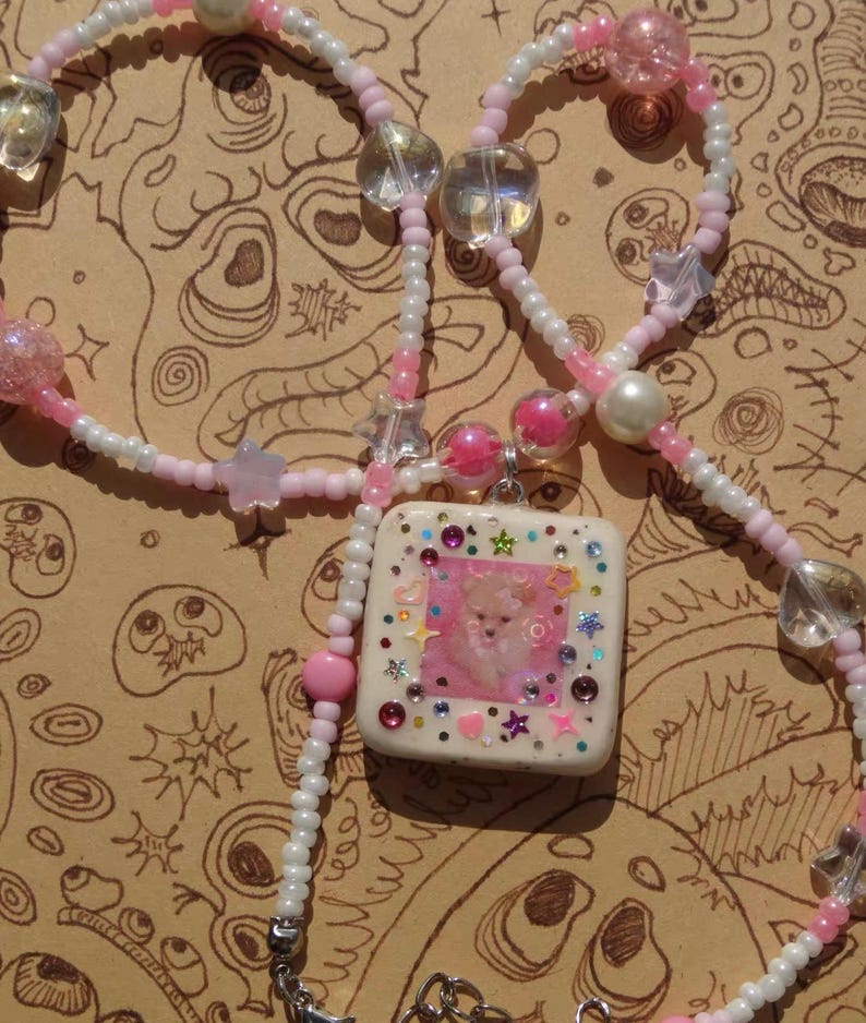 May include: A handcrafted necklace with a square pendant featuring a pink and white design and a small dog image. The necklace has a beaded chain in white and pink, with clear and pink beads. The pendant is decorated with colourful gems and star shapes.