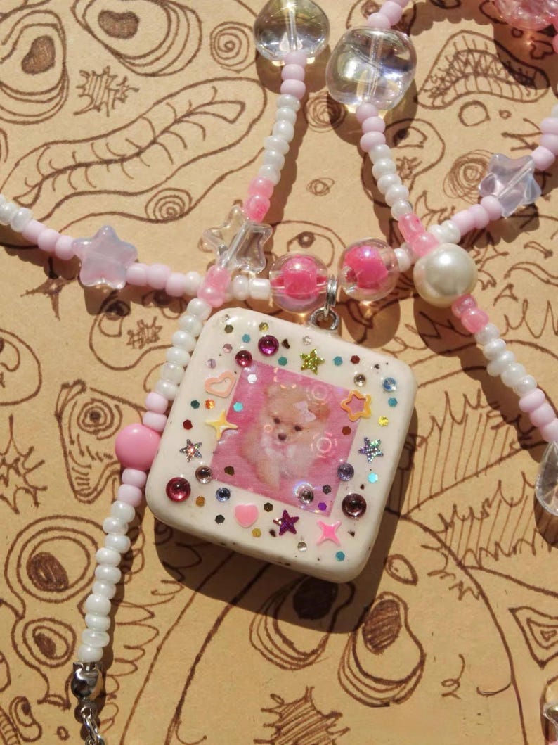 May include: A handcrafted necklace with a square pendant displaying a dog image, embellished with colourful gems and star-shaped details. The necklace features white and pink beaded strands with clear and pink beads.