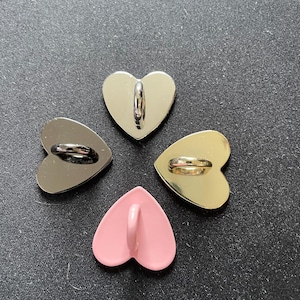 Può includere: Quattro bottoni a forma di cuore in argento, nero, oro e rosa. Ogni bottone ha un anello sul retro per il fissaggio. I bottoni sono disposti in un motivo circolare su uno sfondo scuro.