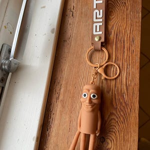 Puede incluir: Un llavero marrón con la palabra "ART" impresa en la correa y una cara sonriente. El llavero presenta una figura de dibujos animados con un cuerpo cilíndrico, dos piernas y pies. El llavero está sujeto a un anillo de metal.