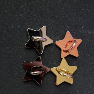 Puede incluir: Cuatro botones metálicos en forma de estrella en plata, oro rosa, metal de cañón y oro. Cada botón tiene un lazo en la parte posterior para coser o sujetar a la tela. Los botones están dispuestos en un cuadrado sobre un fondo oscuro.