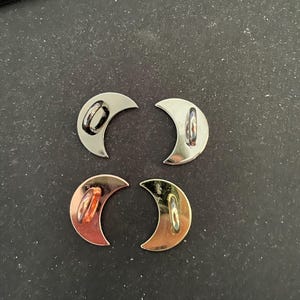 May include: Four crescent moon-shaped metal buttons in silver, gold, and rose gold tones. Each button has a loop on the back for sewing. The buttons are displayed on a dark surface.