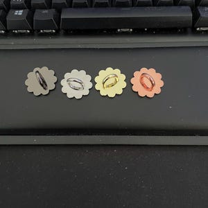 Könnte beinhalten: Vier blumenförmige Metallknöpfe in einer horizontalen Reihe angeordnet. Jeder Knopf verfügt über eine mittige, kreisförmige Öse auf der Vorderseite. Die Knöpfe sind in vier verschiedenen Metallic-Farben gehalten: Mattschwarz, Silber, Gold und Kupfer.