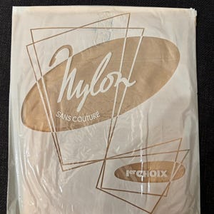 Può includere: Una confezione rettangolare in plastica contenente calze di nylon color beige. La confezione riporta la scritta Nylon, Sans Couture e 1er CHOIX in bianco e marrone, insieme a contorni di triangoli geometrici e una forma ovale sul davanti.