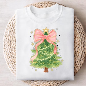 May include: White sweatshirt featuring a watercolor Christmas tree design. The tree is green with a pink bow, gold accents, and string lights. The design includes a gold star on top and small star and dot accents.