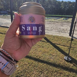 May include: A gold-colored metal tin with a pink and purple label that reads "Sang" in large white letters. The label also includes the text "uplifting & energizing" and a small illustration. The tin is held in a person's hand.