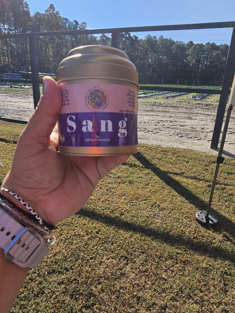 May include: A gold-colored metal tin with a pink and purple label that reads "Sang" in large white letters. The label also includes the text "uplifting & energizing" and a small illustration. The tin is held in a person's hand.