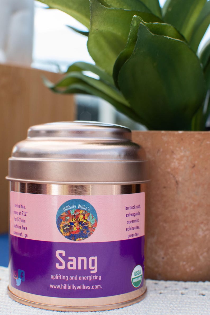 May include: A copper-colored tea tin with a pink and purple label. The label features the text "Sang" and "Hillbilly Willie's" with a colorful cat illustration. The tin is next to a potted plant.