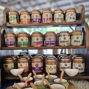 May include: A wooden shelf displays several gold-colored tea tins with various labels, including "Sunshine," "Sang," and "Maia." The tins are arranged on three levels, with small white bowls and wooden spoons in front.