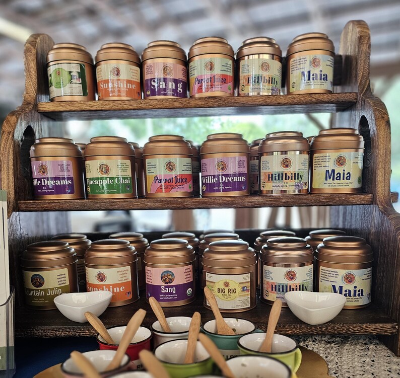 May include: A wooden shelf displays several gold-colored tea tins with various labels, including "Sunshine," "Sang," and "Maia." The tins are arranged on three levels, with small white bowls and wooden spoons in front.