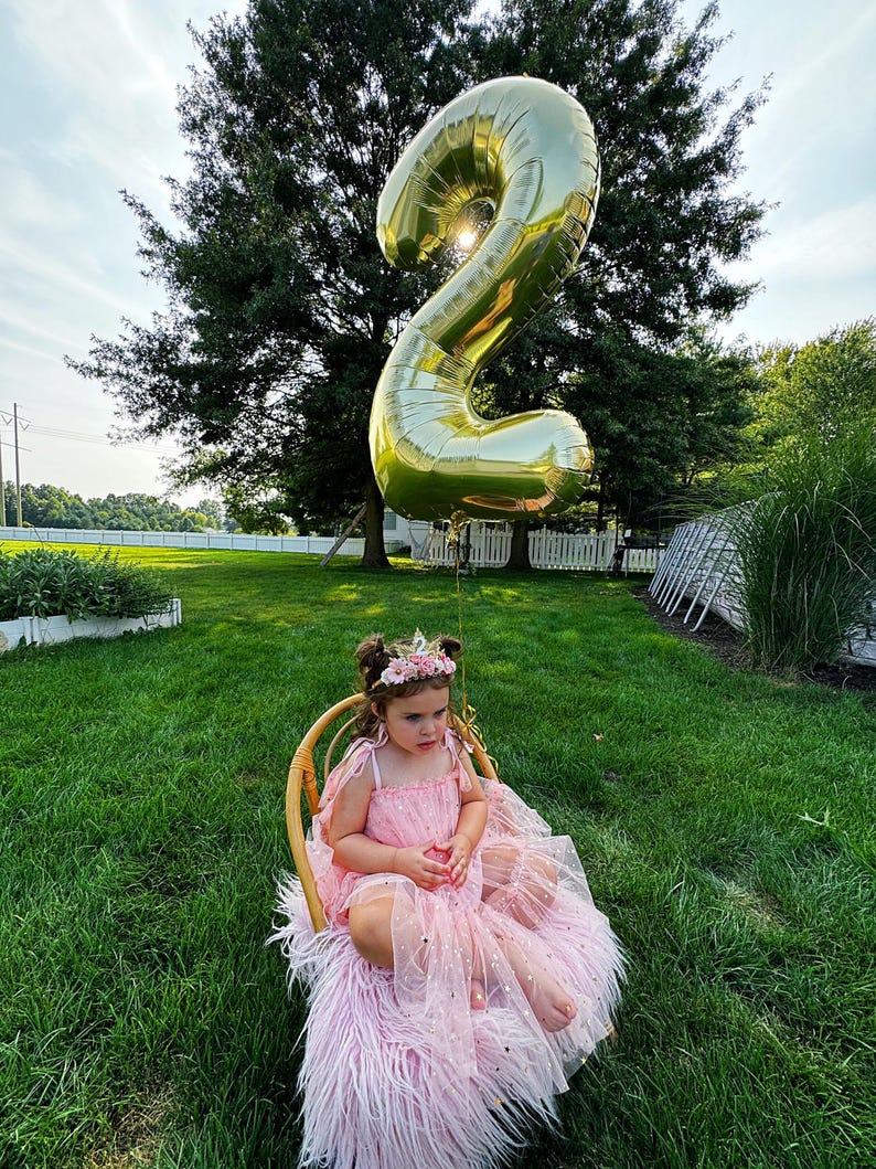 May include: A young girl wearing a pink tulle dress with gold glitter sits in a wicker chair with a large gold number 2 balloon floating above her head.