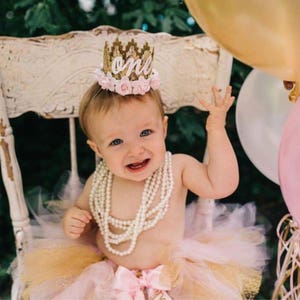 Smiling baby girl wearing a gold lace first birthday crown with pink floral accents, perfect for cake smash photos, milestone photography,  princess-themed birthday parties. A beautiful handcrafted 1st birthday tiara for an unforgettable celebration