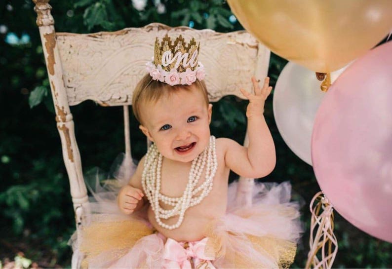 Smiling baby girl wearing a gold lace first birthday crown with pink floral accents, perfect for cake smash photos, milestone photography,  princess-themed birthday parties. A beautiful handcrafted 1st birthday tiara for an unforgettable celebration