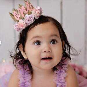 May include: A baby girl wearing a purple dress with gold glitter and a pink and gold crown that says "one" on it.