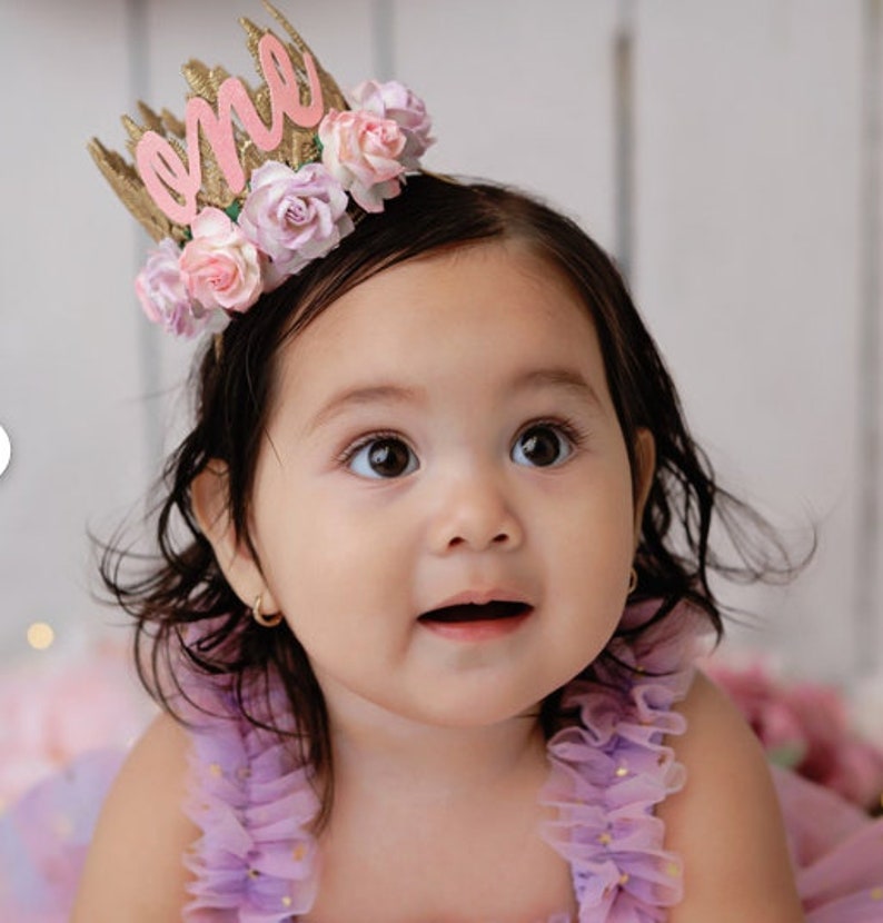 May include: A baby girl wearing a purple dress with gold glitter and a pink and gold crown that says "one" on it.