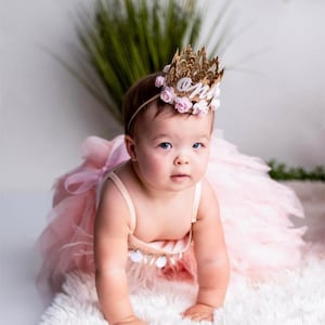 Smiling baby girl wearing a gold lace first birthday crown with pink floral accents, perfect for cake smash photos, milestone photography,  princess-themed birthday parties. A beautiful handcrafted 1st birthday tiara for an unforgettable celebration
