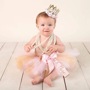 Smiling baby girl wearing a gold lace first birthday crown with pink floral accents, perfect for cake smash photos, milestone photography,  princess-themed birthday parties. A beautiful handcrafted 1st birthday tiara for an unforgettable celebration