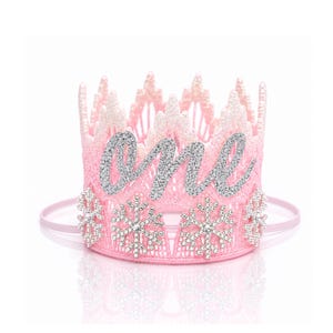 May include: A light pink crown featuring a glittery texture and a pointed top edge. The front displays the word one in silver glitter script, accented by multiple silver snowflake embellishments covered in clear rhinestones.