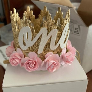 Smiling baby girl wearing a gold lace first birthday crown with pink floral accents, perfect for cake smash photos, milestone photography,  princess-themed birthday parties. A beautiful handcrafted 1st birthday tiara for an unforgettable celebration