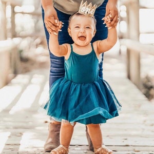 Puede incluir: Una ni&ntilde;a peque&ntilde;a con un vestido de tul azul turquesa y una corona dorada sonr&iacute;e mientras un adulto la sostiene. La ni&ntilde;a est&aacute; de pie en una pasarela de madera.