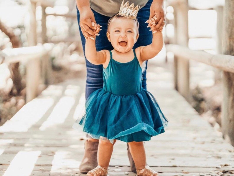 Puede incluir: Una ni&ntilde;a peque&ntilde;a con un vestido de tul azul turquesa y una corona dorada sonr&iacute;e mientras un adulto la sostiene. La ni&ntilde;a est&aacute; de pie en una pasarela de madera.