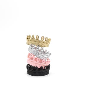 Four stacked crowns in different colors: gold, silver, pink, and black. The crowns are made of a decorative, textured material.
Four tiny lace crowns stacked together in metallic gold, silver, blush pink, and black. Each crown is handcrafted, textured, and made from sculpted lace. This image represents