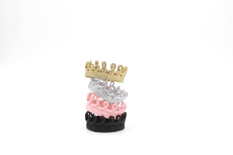 Four stacked crowns in different colors: gold, silver, pink, and black. The crowns are made of a decorative, textured material.
Four tiny lace crowns stacked together in metallic gold, silver, blush pink, and black. Each crown is handcrafted, textured, and made from sculpted lace. This image represents