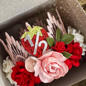 May include: A pink and red flower crown with a strawberry-shaped decoration that says "1 1/2" in silver glitter. The crown is in a white box.