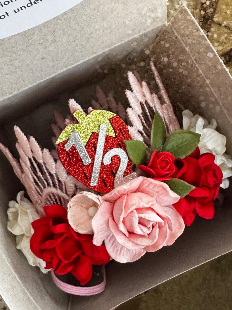 May include: A pink and red flower crown with a strawberry-shaped decoration that says "1 1/2" in silver glitter. The crown is in a white box.