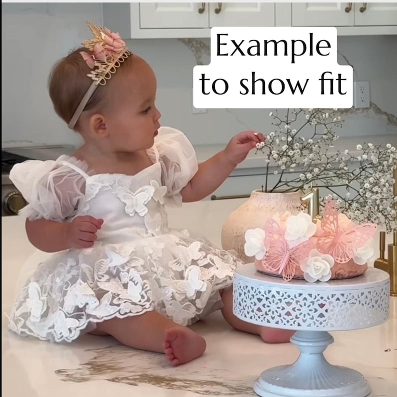 May include: A young child wearing a white tulle dress with butterfly appliques and a gold crown. The child is sitting on a white surface and holding a small white flower. The text "Example to show fit" is visible in the background.