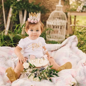 Smiling baby girl wearing a gold lace first birthday crown with pink floral accents, perfect for cake smash photos, milestone photography,  princess-themed birthday parties. A beautiful handcrafted 1st birthday tiara for an unforgettable celebration