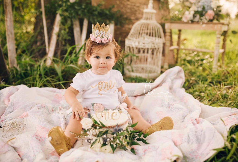 Smiling baby girl wearing a gold lace first birthday crown with pink floral accents, perfect for cake smash photos, milestone photography,  princess-themed birthday parties. A beautiful handcrafted 1st birthday tiara for an unforgettable celebration