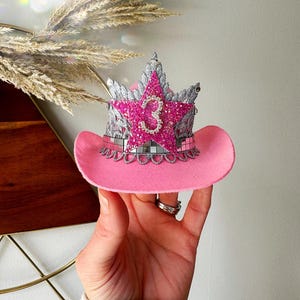 Mini pink cowboy hat birthday crown for baby girls and toddlers, featuring a sparkly silver tiara, hot pink glitter star, and rhinestone number 3 (customizable for any age). Ideal for cowgirl birthday outfits, pink pony parties, or My First Rodeo themes. Shown being held for scale with boho pampas grass in the background. Perfect for first birthdays, cake smashes, dress-up, or western-themed photo shoots.