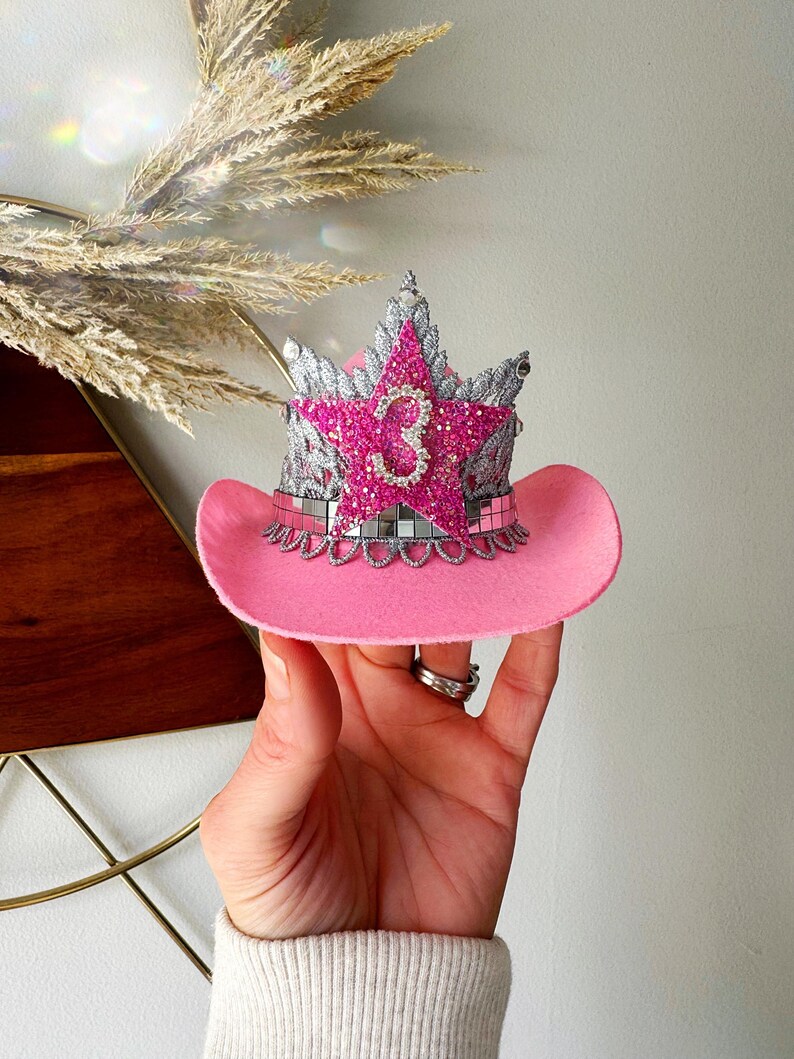 Mini pink cowboy hat birthday crown for baby girls and toddlers, featuring a sparkly silver tiara, hot pink glitter star, and rhinestone number 3 (customizable for any age). Ideal for cowgirl birthday outfits, pink pony parties, or My First Rodeo themes. Shown being held for scale with boho pampas grass in the background. Perfect for first birthdays, cake smashes, dress-up, or western-themed photo shoots.
