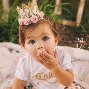 Smiling baby girl wearing a gold lace first birthday crown with pink floral accents, perfect for cake smash photos, milestone photography,  princess-themed birthday parties. A beautiful handcrafted 1st birthday tiara for an unforgettable celebration