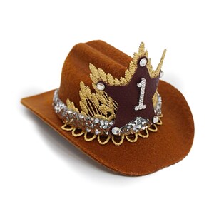Mini brown cowboy hat made of soft felt with a gold faux leather star and metallic trim. The hat features a sparkly number one on the front for a first birthday theme. It’s lightweight, handcrafted, and designed for babies, toddlers, or small pets. Perfect for western birthday parties, rodeo photos, or dress-up costumes. Handmade in the USA by LoveCrush Crowns®.