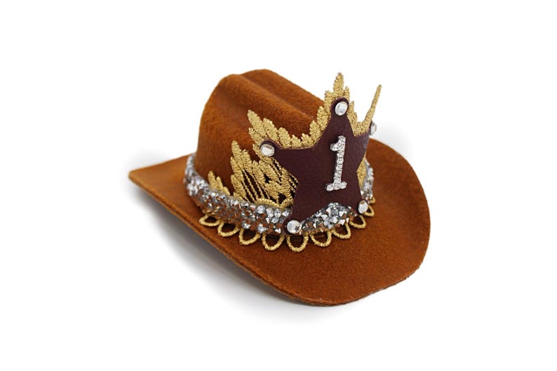 Mini brown cowboy hat made of soft felt with a gold faux leather star and metallic trim. The hat features a sparkly number one on the front for a first birthday theme. It’s lightweight, handcrafted, and designed for babies, toddlers, or small pets. Perfect for western birthday parties, rodeo photos, or dress-up costumes. Handmade in the USA by LoveCrush Crowns®.