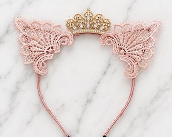 Puede incluir: Una diadema con una corona central de tono dorado adornada con cristales transparentes, flanqueada por dos orejas de gato simétricas de encaje rosa claro. La banda delgada está envuelta en tela rosa a juego con puntas negras.