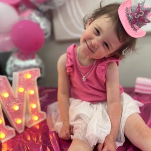 May include: A young child wearing a pink top and a white tutu skirt, smiling at the camera. She is wearing a pink cowboy hat with a silver crown. Pink and silver balloons are in the background, along with illuminated marquee letters.