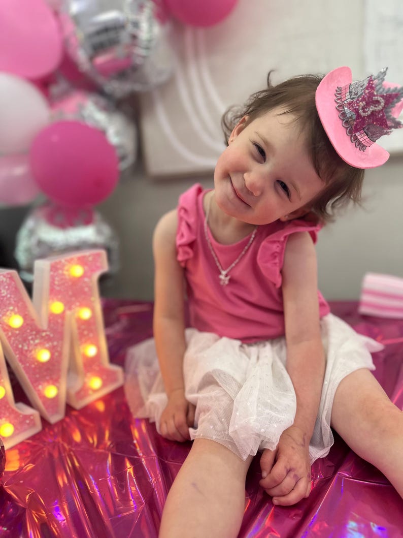 May include: A young child wearing a pink top and a white tutu skirt, smiling at the camera. She is wearing a pink cowboy hat with a silver crown. Pink and silver balloons are in the background, along with illuminated marquee letters.