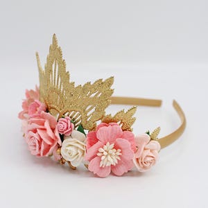 May include: A gold crown headband with pink and white paper flowers. The crown is made of gold lace and has a delicate design. The flowers are arranged around the crown and are accented with gold beads.